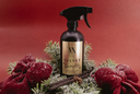 A dark brown spray bottle labeled 'Velvet Sparkle' by The Velvet Noble, styled in a festive holiday wreath with red velvet bows, green pine branches, cinnamon sticks, and faux snow against a solid red background.