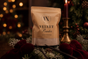 A stand-up pouch of 'The Velvet Noble' Velvet Powder luxury freshener, styled on a rustic table with a lit candle, pine branches, and red velvet bows in a warm, festive holiday setting.