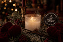 A frosted glass candle holder with a lit candle sits in a holiday wreath with red velvet bows, pinecones, and a black circular sign reading 'THE VELVET NOBLE SILENT NIGHT'.