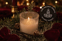 A lit white candle in a textured clear glass holder sits in a holiday wreath with red velvet ribbon, pinecones, evergreen branches, and a round black sign reading 'The Velvet Noble Silent Night'.