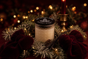 A frosted glass candle jar with a black lid printed with 'Silent Night' and a candle logo, styled in a festive holiday wreath with red velvet bows, pinecones, and warm bokeh lights.