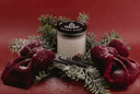 A glass jar candle with a black lid reading 'Silent Night.' is styled with red velvet bows, evergreen branches, and a cinnamon stick on a dark red background.