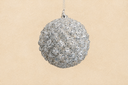 A spherical silver glitter ornament hanging from a thin silver string against a warm beige textured background.