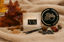 A scented candle in a textured clear glass jar sits on a beige knit blanket with autumn-themed props including fall leaves, cinnamon sticks, vanilla beans, brown sugar, and acorns. The black lid is propped beside the jar, showing the brand 'VIN' and scent 'Vanilla Harvest'.