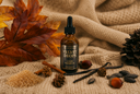 A bottle of Velvet Drops Luxury Warmer Oil centered on a knit fabric background, surrounded by autumn-themed props including leaves, pinecones, cinnamon, vanilla, and acorns.