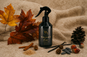 A black spray bottle labeled 'VELVET Home' is styled on a beige fabric surface with autumn leaves, pinecones, cinnamon sticks, vanilla beans, and acorns around it.