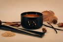 A black cylindrical candle jar with a gold logo plaque reading 'VN The Velvet Noble' sits on a beige surface with autumn-themed props including dried leaves, a pinecone, cinnamon sticks, vanilla beans, and a decorative gold-and-black rod.