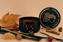 A dark candle tin labeled 'VN The Velvet Noble' with its lid leaning against it, styled with autumn props including a maple leaf, cinnamon sticks, pinecones, a brown sugar pile, and an acorn on a warm beige background.