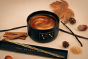 A black jar candle with a swirled amber-brown wax top sits on a beige surface with autumn-themed props including cinnamon sticks, vanilla beans, pinecones, acorns, and a decorative box labeled 'The Velvet Noble'.