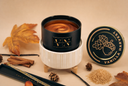 A candle in a matte black jar sits in a white ribbed holder on a beige surface, with the matching lid placed to the right. The scene is styled with autumn leaves, cinnamon sticks, pinecones, vanilla beans, and a small pile of sugar, all under warm lighting.