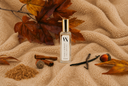 A clear glass perfume bottle with a gold cap and white label reading 'VANILLA HARVEST' and 'Air & Linen Mist', styled on a beige textured blanket with autumn leaves, cinnamon sticks, vanilla bean, and acorns.