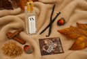 A clear glass perfume bottle with a gold cap and white label reading 'VANILLA HARVEST' and 'Air & Linen Mist', styled on a beige textured blanket with autumn leaves, cinnamon sticks, vanilla bean, and acorns.