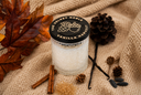 A candle in a textured clear glass jar sits on a beige knit blanket with autumn-themed props including brown leaves, pinecones, acorns, cinnamon sticks, vanilla beans, and a round black label reading 'THE VELVET NOBLE VANILLA HARVEST'.