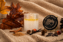 A scented candle in a textured clear glass jar sits on a knitted fabric surface with autumn leaves, cinnamon sticks, pinecones, and acorns around it. A round black label reading 'The Velvet Noble Vanilla Harvest' is visible on the right.