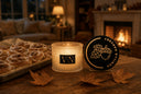 A lit scented candle jar sits on a wooden table beside its round black lid, which reads 'VANILLA HARVEST' with an acorn and leaf illustration. The scene is warmly lit by a fireplace in the background, with cinnamon rolls on a tray and dried leaves on the table.
