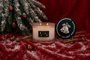 A holiday-themed candle in a clear textured glass jar with two lit wicks, shown next to its black lid on a red velvet surface dusted with faux snow. A decorated Christmas tree branch and cinnamon sticks decorate the background, with the brand name 'Velvet Hollydays' visible on the lid.