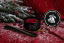 A black wickless candle with red wax sits on a red fabric surface dusted with white faux snow, alongside a matching black lid with gold lettering, cinnamon sticks, red berries, and pine branches with snow in the background. The lid shows the fragrance name 'VELVET HOLLYDAYS'.