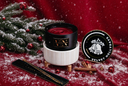 A holiday-themed wickless candle from The Velvet Noble, styled with pine branches, red ornaments, cinnamon sticks, and a black stirring wand and tray on a red fabric background with white faux snow.