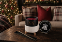 A black and white holiday-themed wickless candle set on a rustic wooden table, featuring a black jar candle with gold lettering, its white ribbed warmer, a matching black lid with a bell illustration, and a black tray. The background shows a cozy living room with a Christmas tree and a couch.