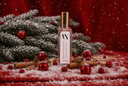 A clear rectangular bottle with a gold cap stands on a red snowy surface with pine branches, red ornaments, cinnamon sticks, and falling snow. The bottle label reads 'VN' and 'Velvet Hollydays'