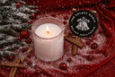 A lit white candle in a textured clear glass holder sits on a red fabric surface dusted with white snow, surrounded by holiday decor including pine branches, red ornaments, cinnamon sticks, and a round black decorative lid reading 'THE VELVET NOBLE VELVET HOLLYDAYS'.