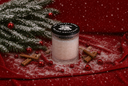 A glass jar candle with a black lid reading 'The Velvet Noble Velvet Hollydays' sits on a red fabric surface dusted with white snow, surrounded by cinnamon sticks, red ornaments, and evergreen branches in a snowy decorative setup.