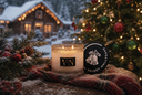 A lit candle in a frosted glass jar sits on a snowy blanket beside a black round tin lid labeled 'VELVET HOLLYDAYS.' A decorated Christmas tree, evergreen branches with red berries, and a snowy cabin with warm lights are softly blurred in the background.