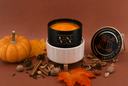 A black and white scented candle labeled 'VN Velvet Pumpkin' is styled with autumn props including a small pumpkin, fall leaves, cinnamon sticks, pinecones, and acorns on a solid terracotta background.
