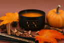 A black candle jar with a gold logo plaque reading 'VN The Velvet Noble' sits on a warm brown surface surrounded by autumn props including orange leaves, a small pumpkin, cinnamon sticks, and pinecones.