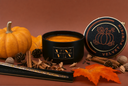 A black candle jar labeled 'VN The Velvet Noble' with orange and shimmering wax, shown beside its matching lid on a warm brown background, styled with a small pumpkin, autumn leaves, cinnamon sticks, pinecones, and nuts.