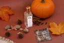 A clear glass bottle with a gold cap and a white label reading 'VN' and 'VELVET PUMPKIN', styled on a warm rust-brown background with a pumpkin, autumn leaves, acorns, cinnamon sticks, and a cream fringed textile.