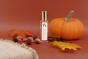 A clear glass bottle with a gold cap and a white label reading 'VN' and 'VELVET PUMPKIN', styled on a warm rust-brown background with a pumpkin, autumn leaves, acorns, cinnamon sticks, and a cream fringed textile.