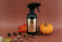 A dark bottle of Velvet Sparkle multipurpose spray with a black trigger sprayer and a gold label, styled with autumn spices, cinnamon sticks, pinecones, a red leaf, and a small pumpkin on a warm brown background.