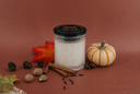 A glass jar candle with white wax and a black lid sits on a warm brown surface, styled with autumn props including a small orange-striped pumpkin, cinnamon sticks, pinecones, walnuts, cloves, and a red leaf.
