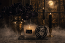 A moody tabletop still life with a lit candle in a textured glass jar, a round black label reading VELVET SPELL, a vase of dark orchids, and two black candles in brass holders against a window view of a moonlit forest.