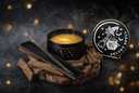 A black tin candle labeled 'VN' sits on a rustic wood slice with a black stirring wand tray, alongside the matching lid printed with a witch hat and broom graphic and the words 'VELVET SPELL.' The scene is styled with wood chips, smoke, and warm golden bokeh lights against a dark background.