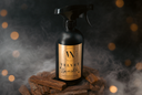 A black trigger spray bottle with a gold label reading 'Velvet Sparkle' by The Velvet Noble, styled on a pile of wood chunks with smoke and warm bokeh lights in the background.