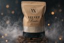 A stand-up pouch of Velvet Powder luxury freshener by The Velvet Noble, displayed on a pile of wood chips with rising smoke against a dark background with warm bokeh lights.