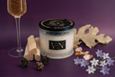 A glass jar candle with a black lid and gold lettering sits on a purple surface, styled with a glass of sparkling drink, wood chips, pinecones, crystals, and small white and lavender flowers. The candle label reads The Velvet Noble & Golden Twilight.