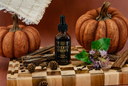 A bottle of Velvet Drops luxury fragrance oil stands on a wooden cutting board surrounded by autumn props including pumpkins, cinnamon sticks, pinecones, and a purple flower, against a warm brown background.