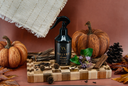 A black spray bottle of Velvet Home luxury air & linen mist by The Velvet Noble, styled on a wooden board with pumpkins, pinecones, and a rustic woven textile against a warm brown background.