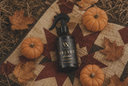 A black spray bottle labeled 'VELVET HOME' lies on a patterned autumn quilt surrounded by small pumpkins and dried leaves.