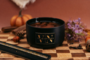 A black cylindrical candle jar with a gold 'VN' logo and 'The Velvet Noble' text, filled with a warm gold glittered wax, styled on a wooden checkerboard with pumpkins, purple flowers, and spices against a terracotta background.