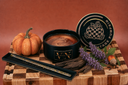A black candle tin labeled 'VINTAGE PUMPKIN' sits on a wooden checkered board with a small pumpkin, bark pieces, and a round branded lid against a warm brown background.