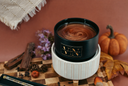 A black ceramic candle jar with a gold logo, filled with a gold glittered swirled wax, styled on a wooden board with autumn props including a small pumpkin, leaves, cinnamon sticks, and purple flowers against a terracotta background.