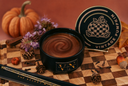 A black jar candle with a swirled gold glitter wax top sits on a wooden checkerboard surface, with its matching lid leaning behind it. The scene includes a pumpkin, flowers, pinecones, cinnamon sticks, acorns, and a decorative pen, all styled against a warm terracotta background.