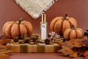A clear rectangular bottle with a gold cap stands centered on a wooden board, surrounded by decorative autumn props including small pumpkins, leaves, pinecones, and a small purple flower. The background is a warm terracotta wall with a cream textile hanging at the top.
