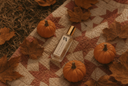 A clear rectangular bottle with a gold cap lies on a quilted blanket surrounded by autumn leaves and small pumpkins.