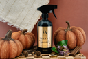 A black spray bottle labeled 'Velvet Sparkle' multipurpose cleaner, styled on a wooden board with decorative pumpkins, cinnamon sticks, pinecones, and purple flowers against a terracotta background.