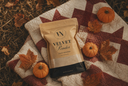 A stand-up pouch of Velvet Powder by The Velvet Noble lies on a patterned quilt outdoors, surrounded by small pumpkins and autumn leaves.